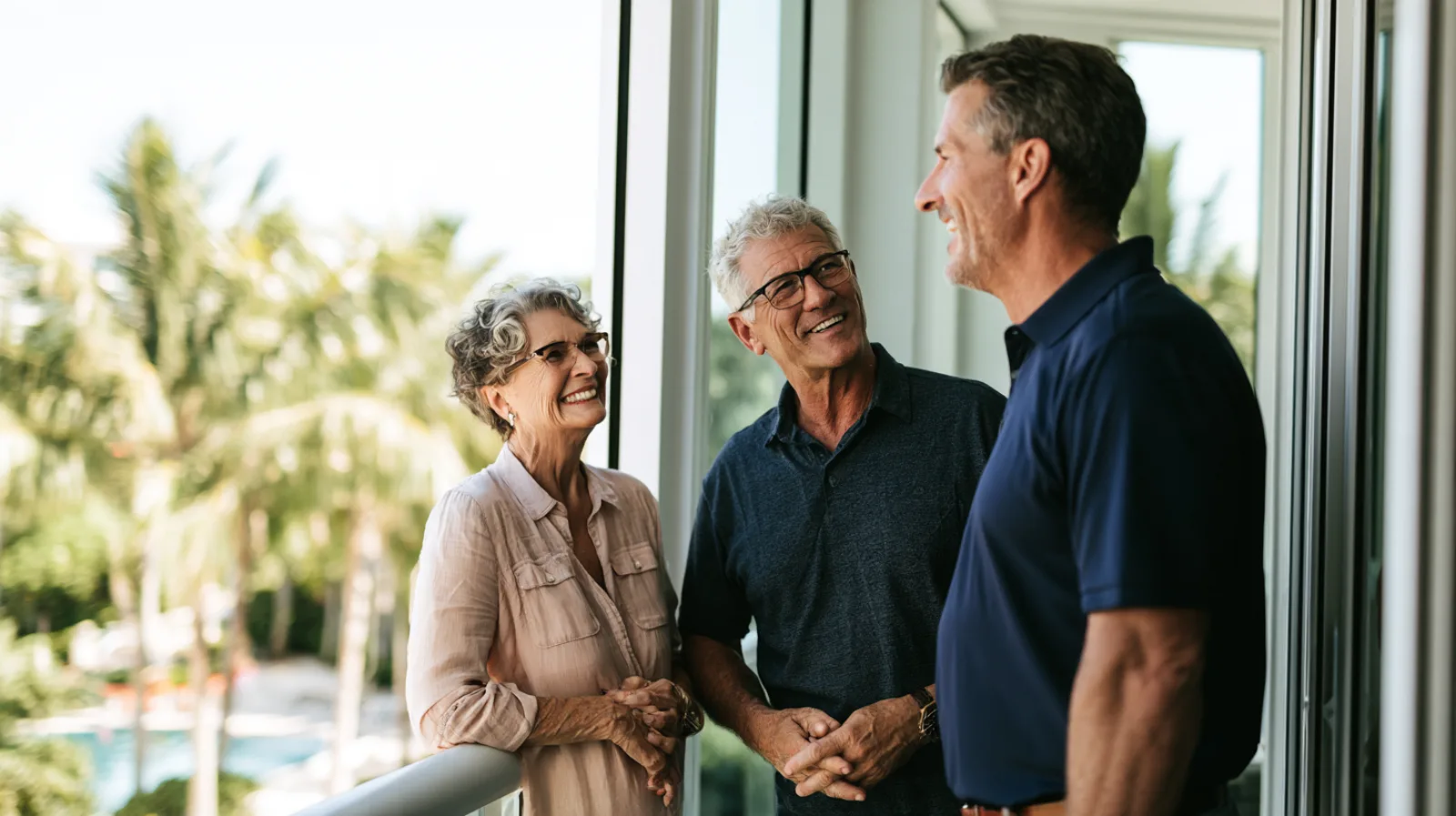 A smiling retired couple talks with a technician in a navy polo on the balcony of their bright South Florida condo, with palm trees and a pool below.