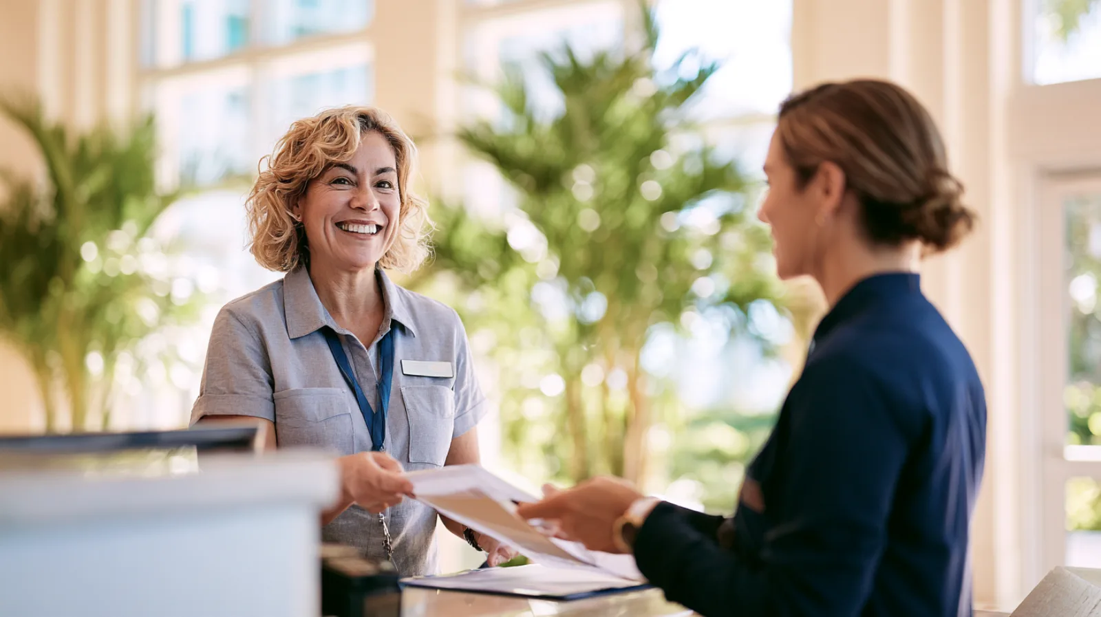 A smiling building manager takes a folder of paperwork from a condo owner across the front desk of a bright lobby with potted palms.