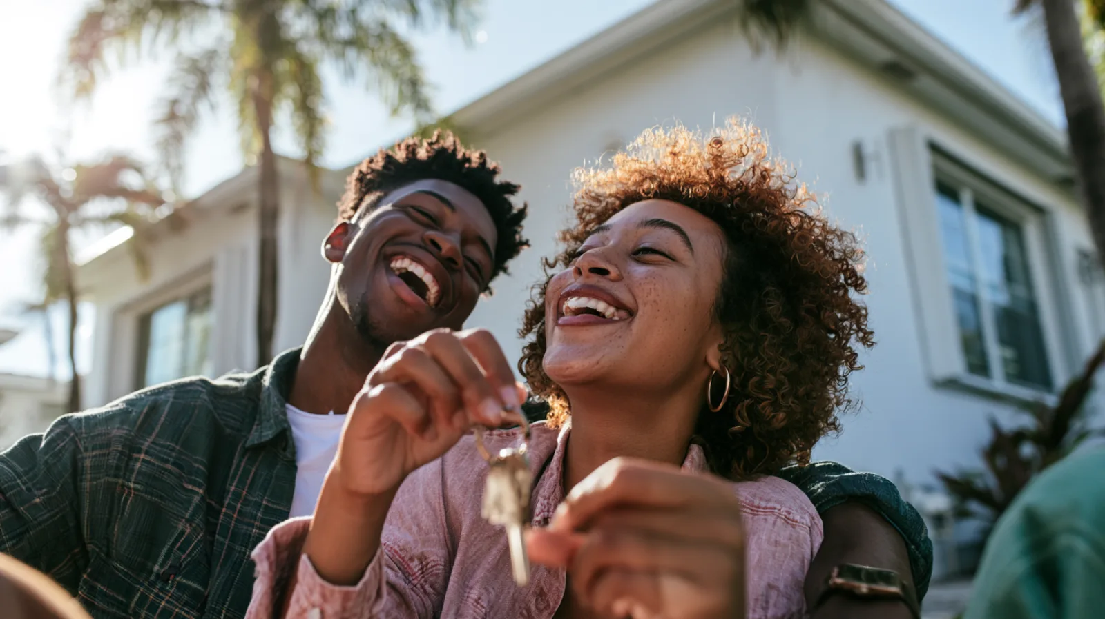 A laughing young couple holds up the keys to their new home outside a white single-story house with palm trees.