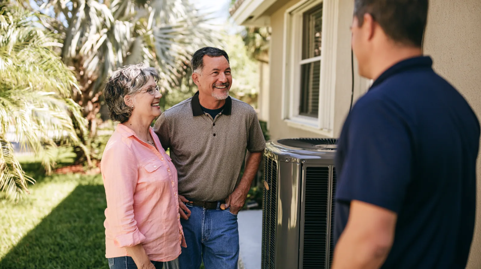 A smiling couple stands beside their new outdoor air conditioner at the side of their South Florida home, talking with a technician in a navy polo.