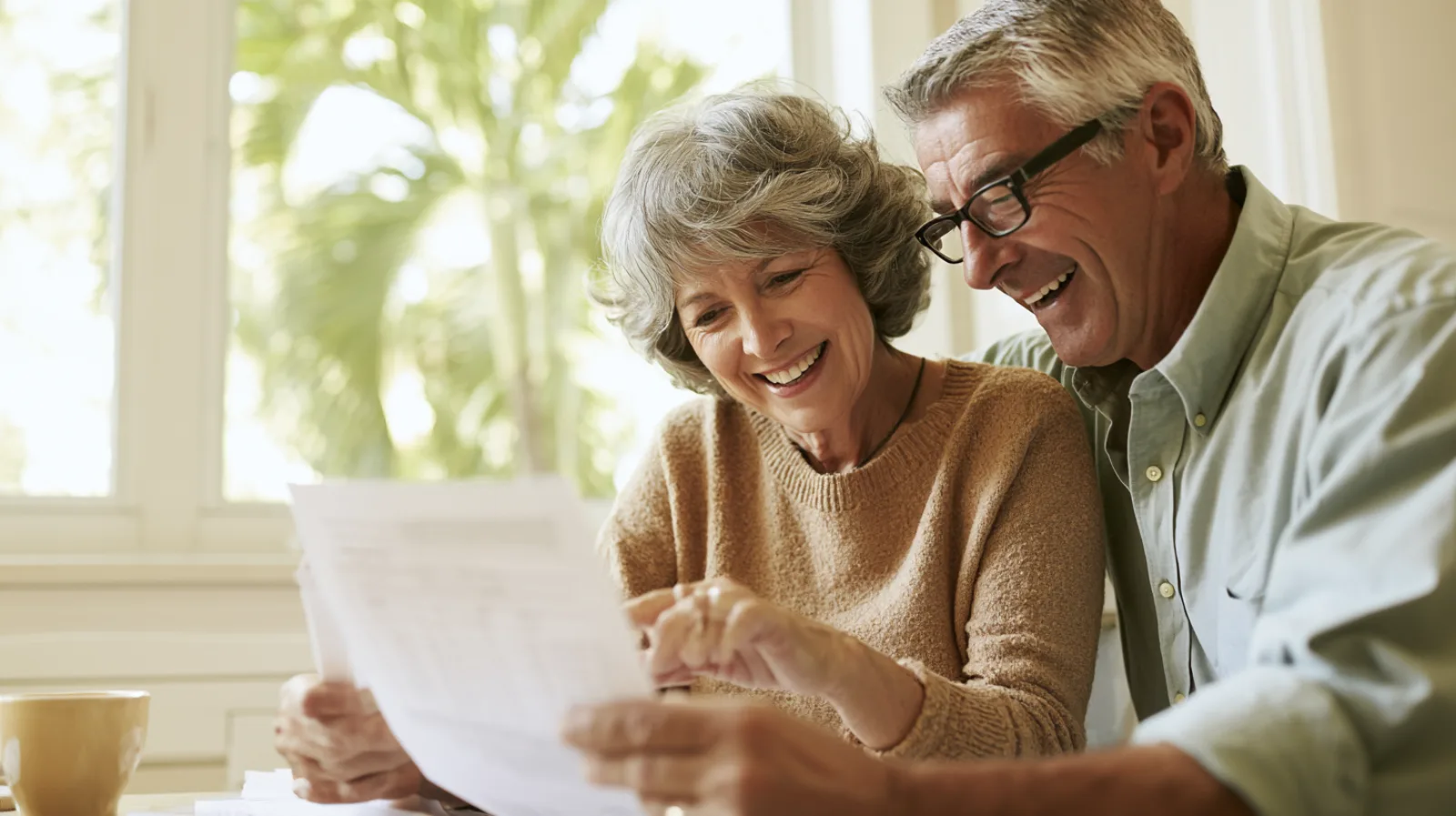 A smiling couple in their sixties read a printed AC replacement quote together at a bright kitchen table, with a palm tree outside the window.