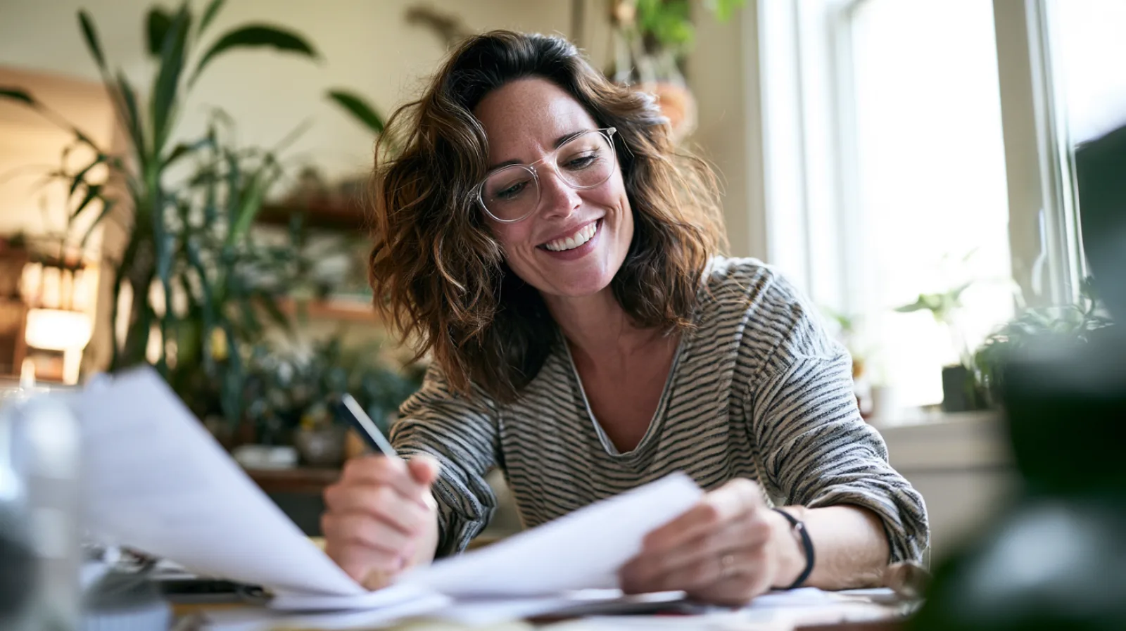A smiling woman with glasses compares two printed quotes side by side at a desk in a bright room full of plants.