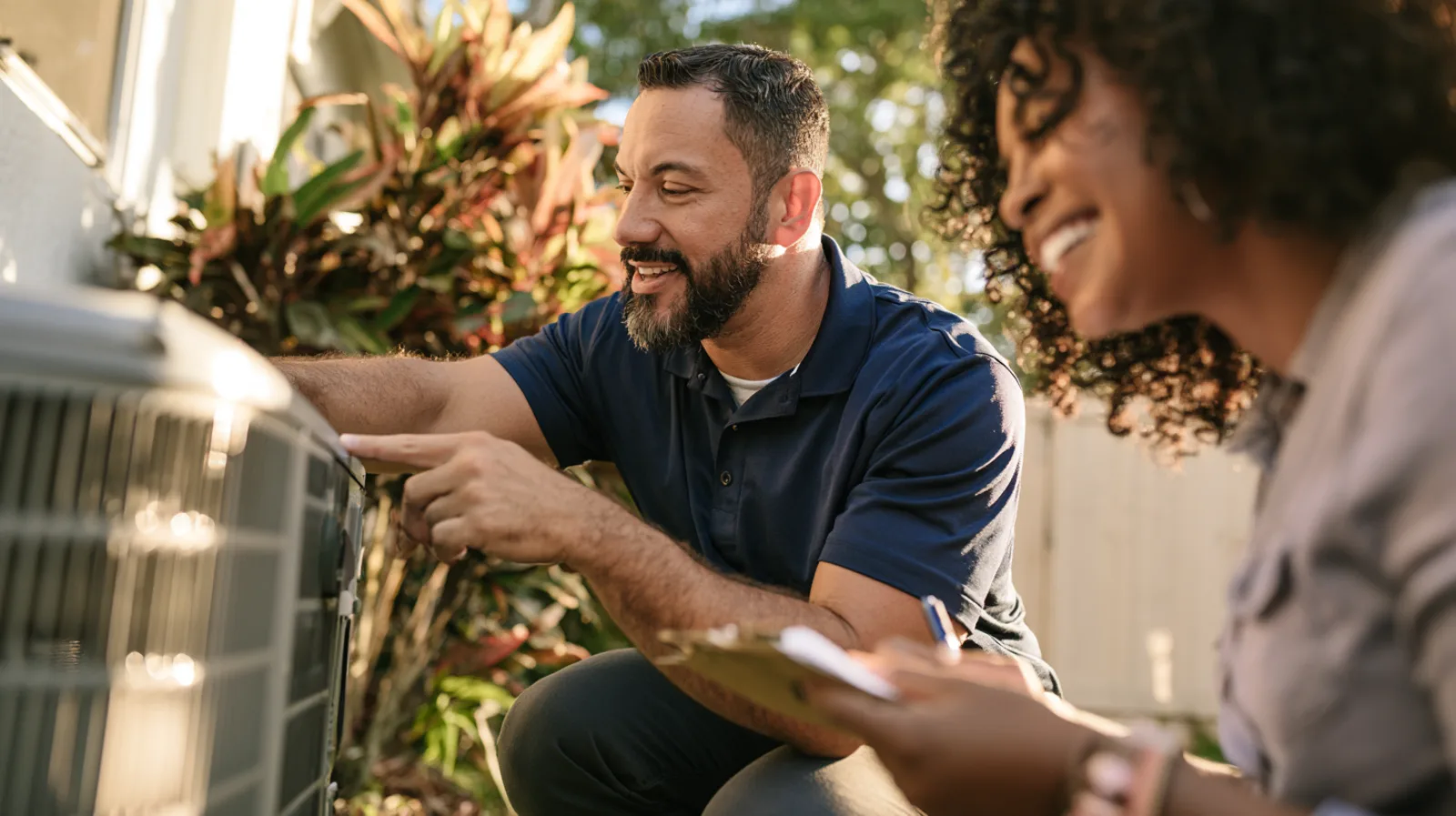 A technician in a navy polo crouches beside an outdoor air conditioner and points at the unit while a smiling homeowner takes notes on a clipboard.