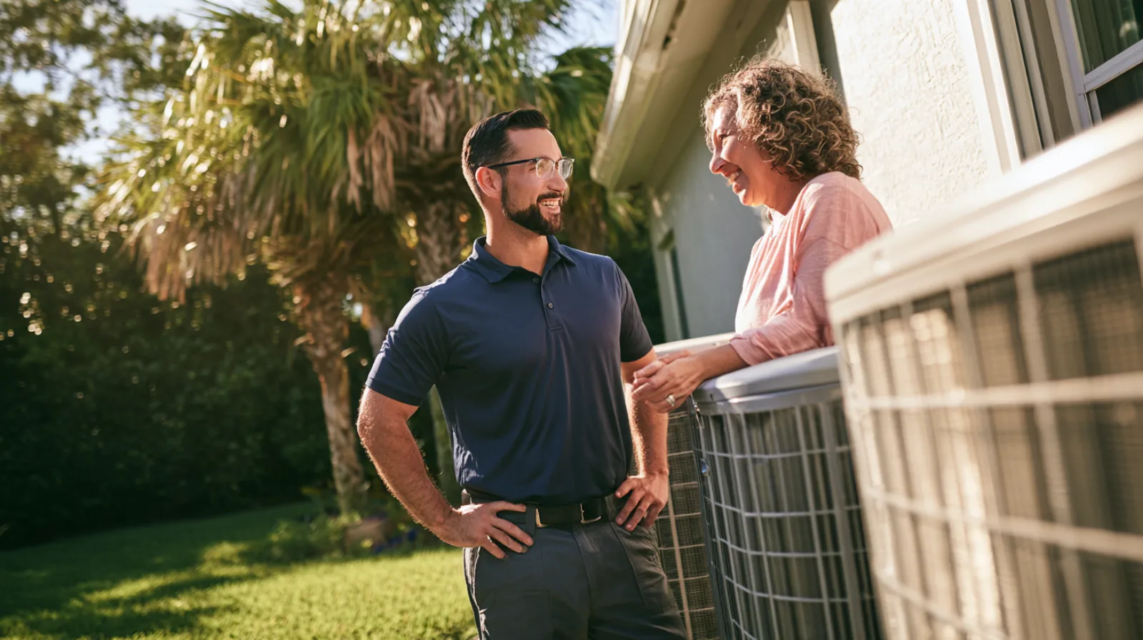 A technician in a navy polo talks with a smiling homeowner beside two outdoor air conditioner units at the side of a Florida house, with palm trees behind them.