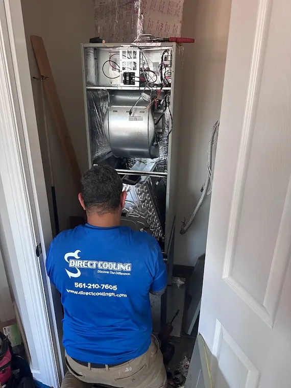 A Direct Cooling technician works on an air handler with its panels off, blower and coil exposed, in a closet. Direct Cooling technician working inside an open air handler in a closet