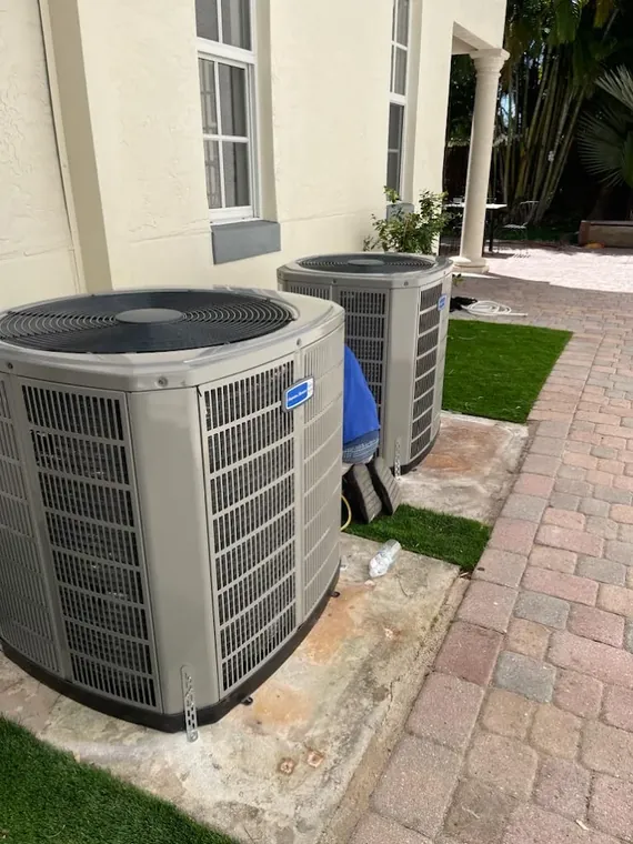 Two American Standard outdoor condensers on pads beside a residence, with a technician working between them. Two American Standard AC condensers installed side by side beside a home