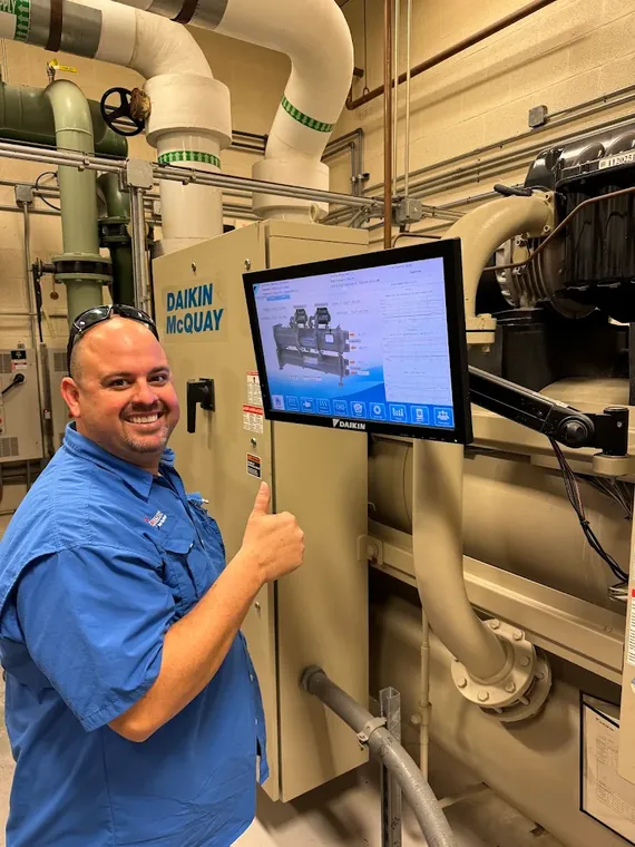 A Direct Cooling technician in a mechanical room beside a Daikin McQuay chiller and its touchscreen controls. Direct Cooling technician giving a thumbs up beside a Daikin chiller control screen