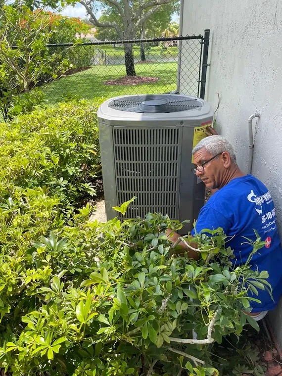 Direct Cooling technician servicing an outdoor AC condenser behind landscaping