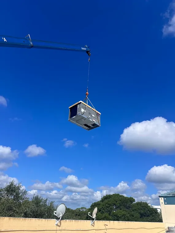 A crane hoists a new packaged rooftop AC unit onto a commercial building during a Direct Cooling installation. Crane lifting a new rooftop package air conditioning unit against a blue sky