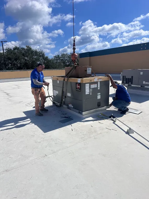 Direct Cooling technicians guide a new Rheem commercial rooftop unit into place on the roof curb as the crane lowers it. Two Direct Cooling technicians guiding a Rheem commercial rooftop unit onto its curb