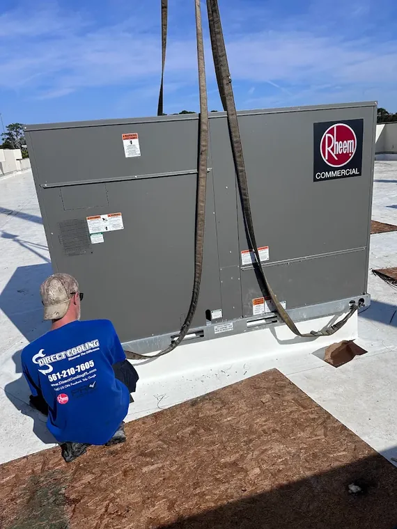 A Direct Cooling technician checks a Rheem commercial rooftop unit still rigged in its lifting straps on the roof. Direct Cooling technician beside a Rheem commercial unit still in its lifting straps